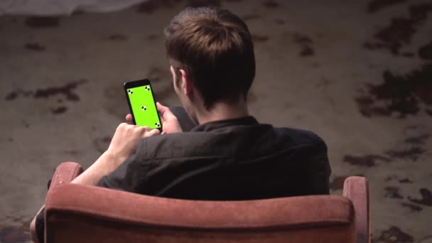 Bovenaanzicht van jonge man met bruin haar zittend in stoel en tikken op de iPhone groen scherm in donkere kamer. Stock footage. Chroma Key — Stockvideo