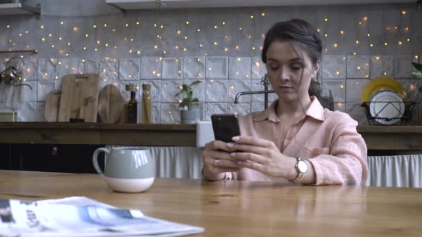 Hermosa mujer joven con el pelo castaño en camisa rosa sentado en la cocina en la mesa de madera y el uso de teléfono inteligente. Imágenes de archivo. Interior de la casa decorada moderna en el fondo — Vídeos de Stock