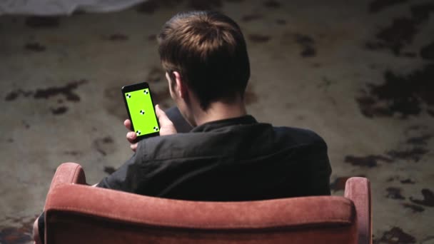 Aantrekkelijke jonge man met bruin haar zittend in comfortabele stoel met wit kussen en glijden op iPhone groen scherm in donkere kamer. Stock footage. Chroma Key — Stockvideo