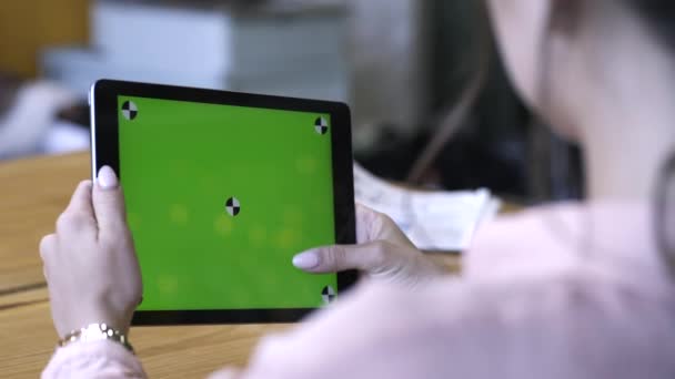 Vista desde el hombro de una mujer joven con camisa rosa sentada en la mesa y deslizándose en su tableta en la pantalla verde croma key. Imágenes de archivo. Pantalla de clave de croma para la colocación de su propio contenido . — Vídeos de Stock