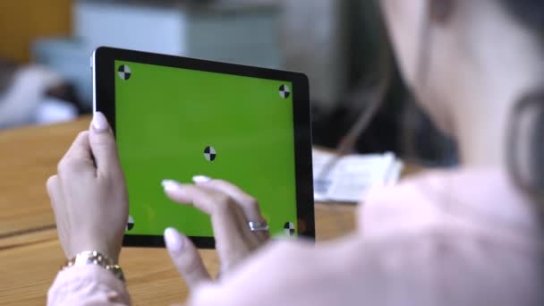 茶色の髪をした若い女性がテーブルに座り、クロマキーグリーンスクリーンでタブレットを滑らせる。ストック映像。独自のコンテンツを配置するためのクロマキー画面. — ストック動画