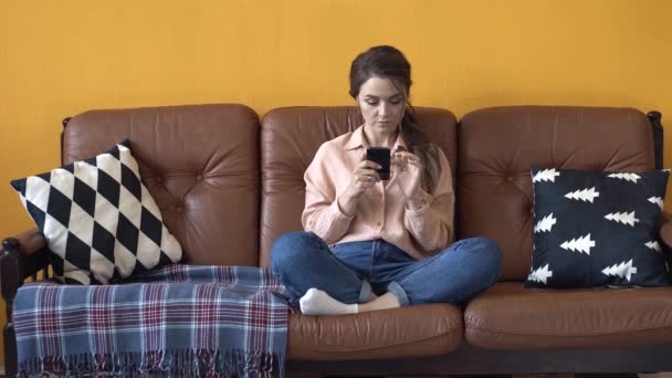 Mujer joven feliz con el pelo oscuro en camisa rosa y vaqueros sentados en el sofá marrón con almohadas y escribiendo en su teléfono inteligente. Imágenes de archivo. Mujer relajándose en casa — Vídeos de Stock