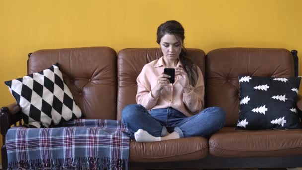 Lycklig ung kvinna med mörkt hår i rosa skjorta och jeans sitter på den bruna soffan med kuddar och skriva på hennes smartphone. Stock film. Kvinna avkopplande hemma — Stockvideo