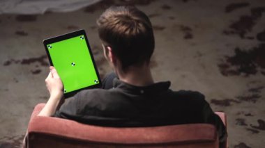 Siyah gömlekli genç adamın arkasından sandalyede oturan ve izleme işaretleri ile dijital tablet bilgisayarın yeşil mock-up ekran dokunarak görünümü. Stok görüntüleri. Renk anahtar ekranı