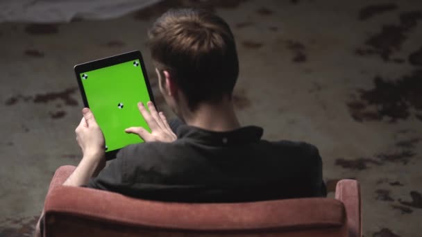 Utsikt från baksidan av ung man i svart skjorta sitter i stolen och knacka på grön mock-up skärm av digital Tablet PC med spårnings märken. Stock film. Chroma nyckel skärm — Stockvideo