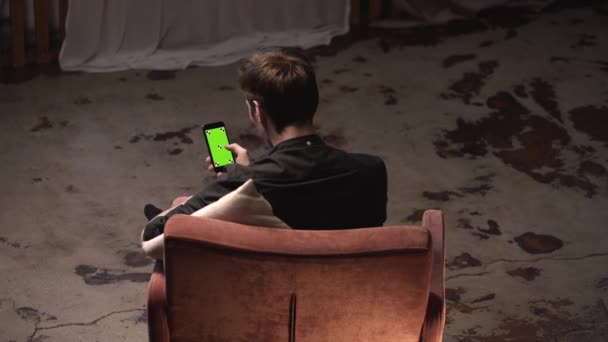 Aantrekkelijke jonge man met bruin haar zittend in comfortabele stoel met wit kussen en glijden op iPhone groen scherm in donkere kamer. Stock footage. Chroma Key — Stockvideo