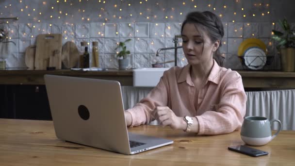 Härlig ung kvinna sitter i köket på träbord och skriva på sin laptop med koppen te eller kaffe. Stock film. Modernt inrett Heminredning på bakgrunden — Stockvideo