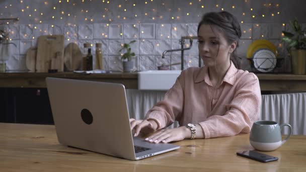 Härlig ung kvinna sitter i köket på träbord och skriva på sin laptop med koppen te eller kaffe. Stock film. Modernt inrett Heminredning på bakgrunden — Stockvideo
