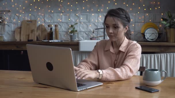 Close-up van aantrekkelijke jonge jonge vrouw werken op laptop computer terwijl zittend op een keuken. Stock footage. Modern ingericht huis interieur op de achtergrond — Stockvideo