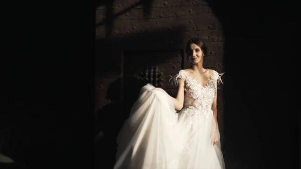 Vista de cerca de la joven novia hermosa en un vestido de novia blanco de moda de pie y sonriendo cerca de la vieja pared oscura y puertas. Acción. Una boda de cuentos —  Fotos de Stock