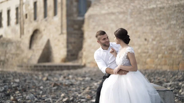 Close-up of beautiful fairytale newlywed couple sitting on the bench and hugging at rocky beach near old medieval castle in France. Action. A storybook wedding — Stock Photo, Image