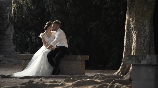 Όμορφο παραμυθένιο νεόνυμφο ζευγάρι κάθεται στο πέτρινο παγκάκι και φιλιέται στο πάρκο. Πάμε. Ο κομψός γαμπρός αγκαλιάζει την όμορφη νύφη στον κήπο. — Φωτογραφία Αρχείου