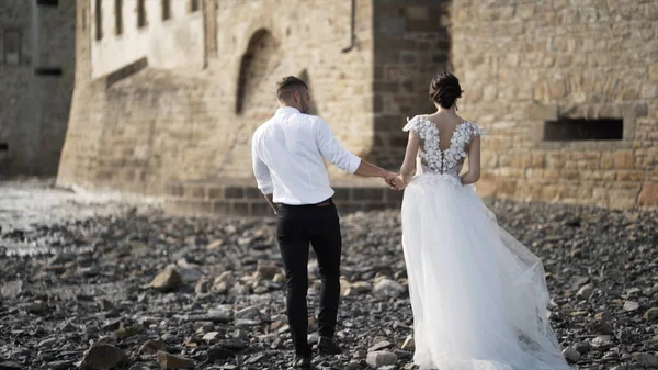 View from the back of beautiful fairytale newlywed couple walking on the rocky beach near old medieval castle in France. Action. A storybook wedding