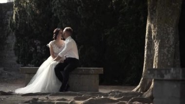 Güzel peri masalı yeni evli çift taş bankta oturan ve parkta öpüşme. Eylem. Şık damat bahçede güzel gelin kucaklıyor.