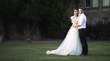 Yeşil çim en iyi şekilde duran ve ağaçların ve büyük gri binanın yakınında sarılan inanılmaz beyaz elbiseli damat ve gelinin güzel düğün çifti. Eylem. Bir hikaye kitabı düğün