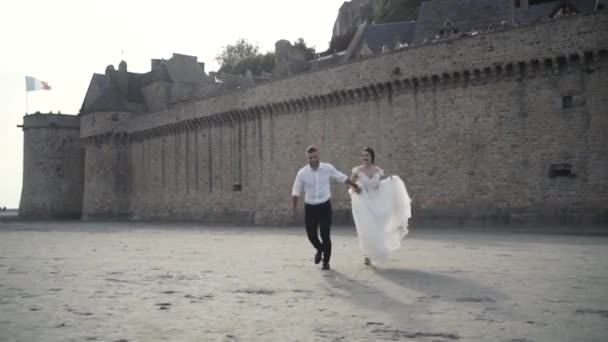 Close-up view of beautiful fairytale newlywed couple fleeing on the sand near old medieval castle in France against blue sky. Action. A storybook wedding — Stock Video