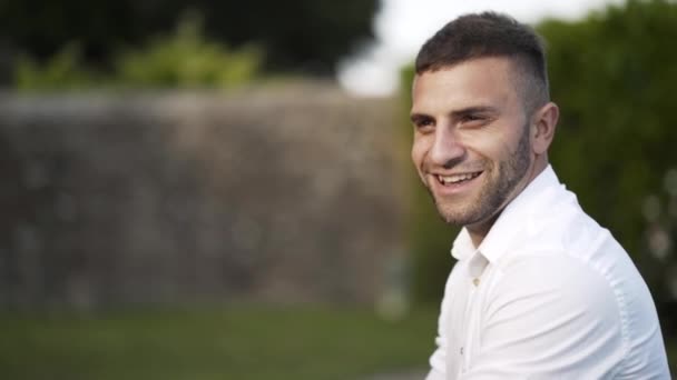 Vista de perto do belo noivo com cabelo castanho e barba em camisa branca olhando para a câmera e sorrindo no parque. Acção. Vídeo de casamento — Vídeo de Stock