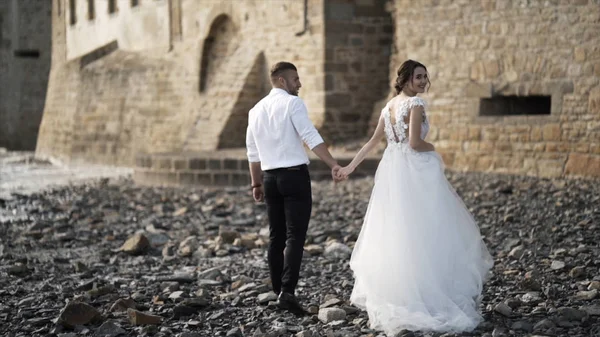 View from the back of beautiful fairytale newlywed couple walking on the rocky beach near old medieval castle in France. Action. A storybook wedding