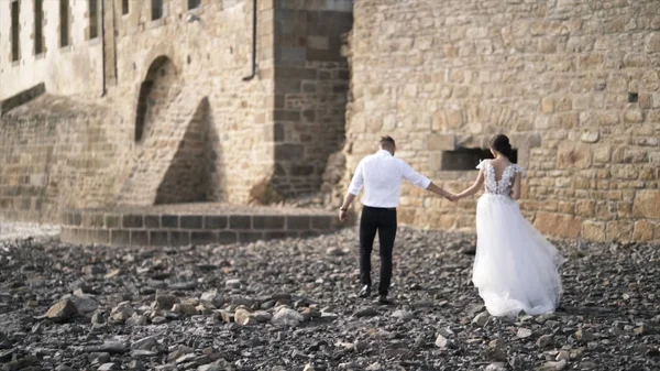 View from the back of beautiful fairytale newlywed couple walking on the rocky beach near old medieval castle in France. Action. A storybook wedding