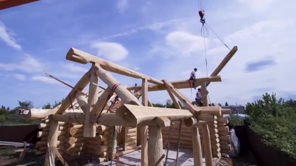 Los trabajadores en la construcción de la casa de campo con grúa. Clip. Construcción de casa de madera de campo. Constructores en diseño de madera llevan a casa vigas de grúa en el fondo del cielo azul — Vídeo de stock