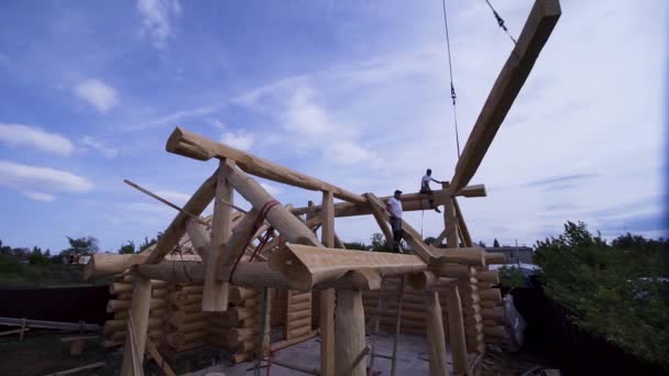 Workers at construction of cottage with crane. Clip. Construction of country wooden house. Builders in wood design take home beams of crane on background of blue sky — Stock Video