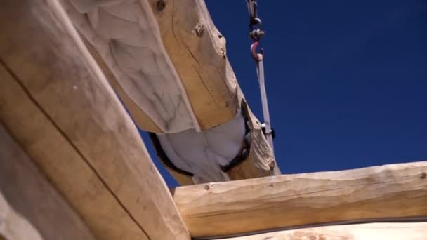 Close-up of wooden house and lifting crane with timber. Clip. Wooden house under construction on background of crane with log. Wooden structure of house on background of blue sky — Stock Footage Close-up of wooden house and lifting crane with timber. Clip. Wooden house under construction on background of crane with log. Wooden structure of house on background of blue sky Stock Video