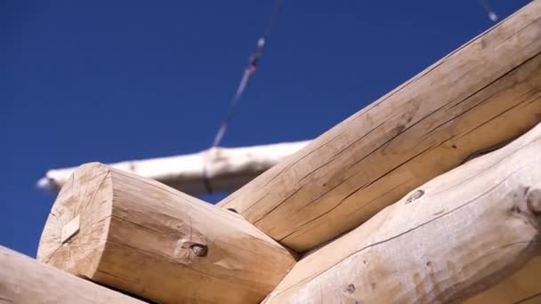 Крупный план деревянного дома и подъемный кран с древесиной. Клип. Деревянный дом строится на фоне крана с бревном. Деревянная структура дома на фоне голубого неба — стоковое видео
