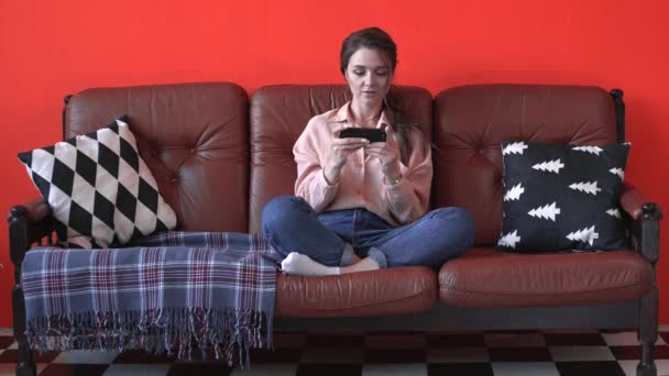 Close-up view of young pretty woman in pink shirt and jeans sitting on the brown couch with pillows and play mobile game by phone. Stock footage. Feeling of happy and relax at home — Stock Video