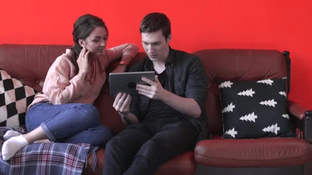 Närbild av glada unga par sitter på den bruna soffan hemma och tittar på tablettskärmen. Lagerbilder. Man och kvinna kopplar av i soffan, glad helg — Stockvideo