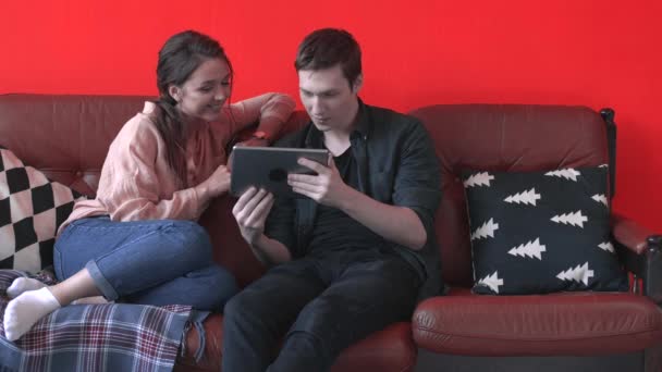 Vista de cerca de la feliz pareja joven sentada en el sofá marrón en casa y mirando en la pantalla de la tableta. Imágenes de archivo. Hombre y mujer relajándose en el sofá, feliz fin de semana — Vídeo de stock