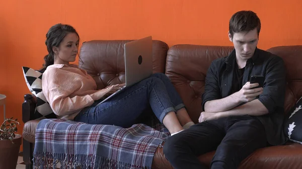 Nahaufnahme eines jungen Mannes und einer jungen Frau, die zusammen auf einer braunen Ledercouch zu Hause sitzen, während sie Laptop und Smartphone benutzen. Archivmaterial. junges Paar entspannt zu Hause. — Stockfoto