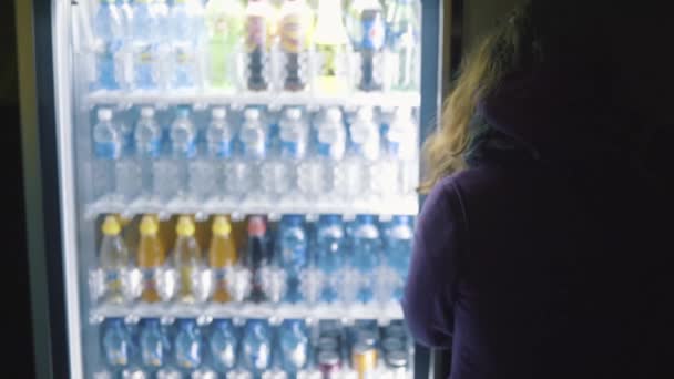 Вид с задней стороны молодой женщины в фиолетовом пальто, выбирающей напиток из торгового автомата. Запись. Торговый автомат по продаже воды в бутылках — стоковое видео