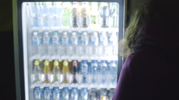 Вид с задней стороны молодой женщины в фиолетовом пальто, выбирающей напиток из торгового автомата. Запись. Торговый автомат по продаже воды в бутылках — стоковое видео