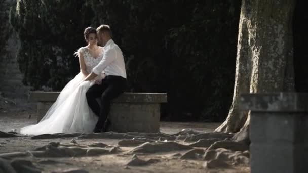 Hermosa pareja recién casada de cuento de hadas sentada en el banco de piedra y besándose en el parque. Acción. El novio con estilo abraza a la novia encantadora en el jardín . — Vídeos de Stock