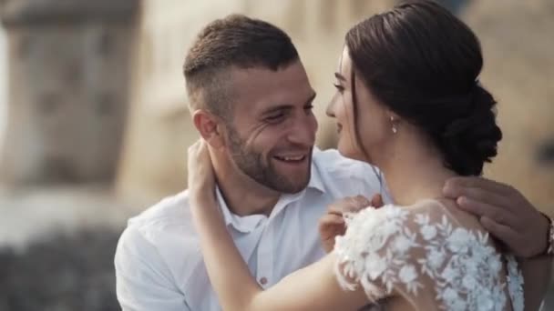 Close-up of beautiful fairytale bride in a fashionable white wedding dress and groom sitting in the bench, smiling and kissing. Action. A storybook wedding — Stock Video
