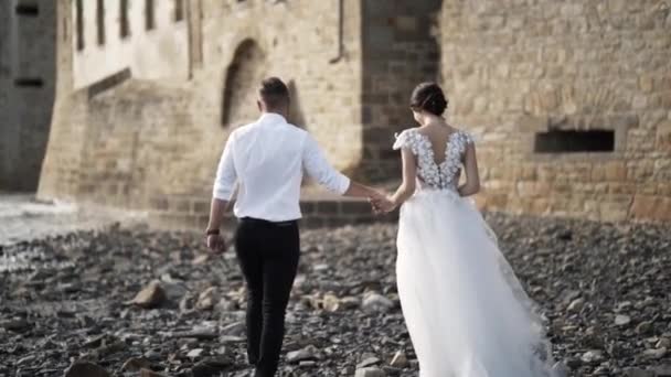 Uitzicht vanaf de achterkant van het prachtige sprookje jonggehuwde paar wandelen op het rotsachtige strand in de buurt van oude middeleeuwse kasteel in Frankrijk. Actie. Een Storybook bruiloft — Stockvideo