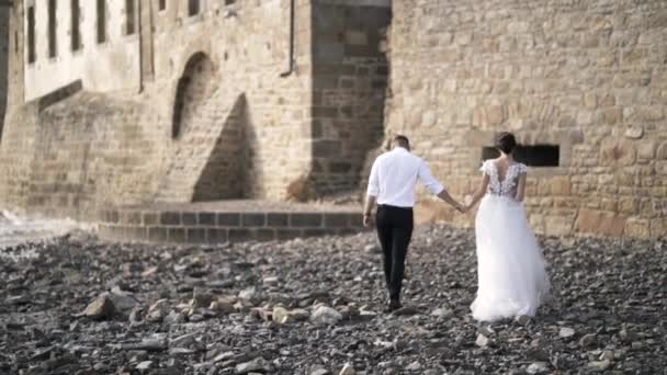 Vue de l'arrière d'un beau couple de contes de fées marchant sur la plage rocheuse près du vieux château médiéval en France. L'action. Un mariage de livre d'histoires — Video