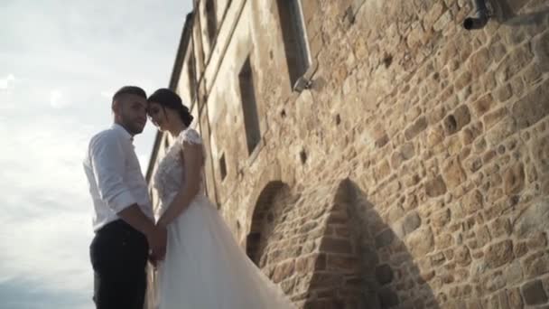 Mooie Fairytale Newlywed paar poseren in de buurt van oude kasteel muur. Actie. De stijlvolle bruidegom houden handen van de mooie bruid. — Stockvideo