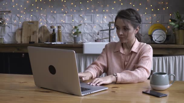 Närbild av attraktiva ung ung kvinna som arbetar på bärbar dator när du sitter på ett kök. Stock film. Modernt inrett Heminredning på bakgrunden — Stockvideo