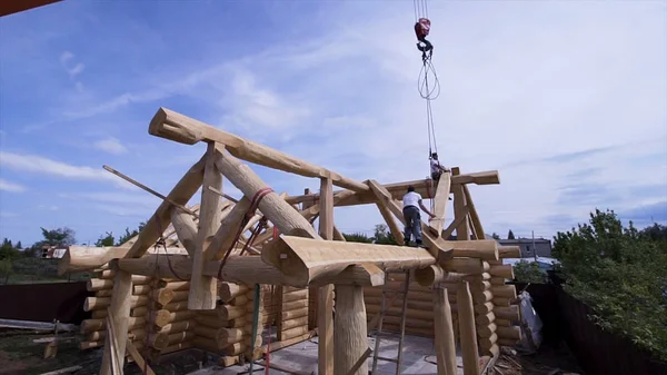 Workers at construction of cottage with crane. Clip. Construction of country wooden house. Builders in wood design take home beams of crane on background of blue sky