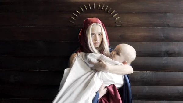 头戴头巾的妇女，抱着婴儿的母亲，靠着木制墙壁站着，带着子弹、宗教和战争的概念。 镜头。 圣母玛利亚和耶稣的形象. — 图库照片