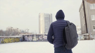 Koyu mavi ceketli ve sırt çantalı bir adam şehrin sokaklarına ve duvar yazılarına bakıyor. Başla. Gri gökyüzü arka planında bir kış şehrinde bir öğrencinin arkası..