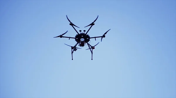 Quadcopter dron vznášející se na jasně modré obloze, video a fotografování koncepce. Klip. Malý černý quadrotor s digitálním fotoaparátem a otočnými lopatkami, spodní pohled. — Stock fotografie