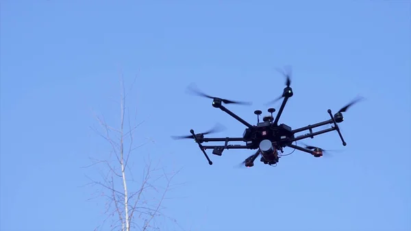 Black camera drone in flight with visible propelers movement and flashing light on blue sky background. Clip. Small quadcopter hovering against clear blue sky, slow motion.