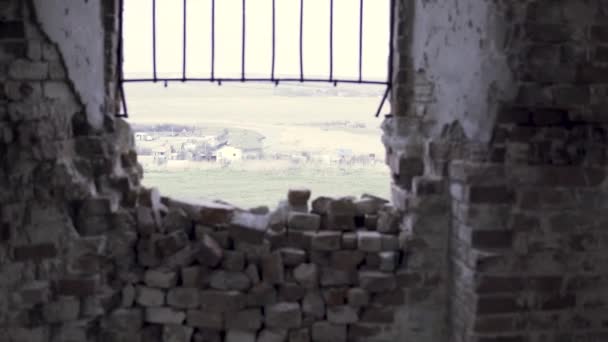 Vista do campo verde e uma aldeia do escuro destruiu o velho edifício de tijolos através da janela. Filmagem. Uma parede rachada velha e uma armação de janela que olha um prado verde e casas de madeira . — Vídeo de Stock