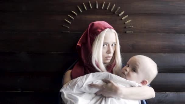 Közelkép egy ijedt fiatal nőről, aki piros csuklyát visel, fehér lepedőben tartva egy babát, fából készült fali háttérrel. Felvétel. Szűz Mária és Jézus képe, vallási fogalom. — Stock videók