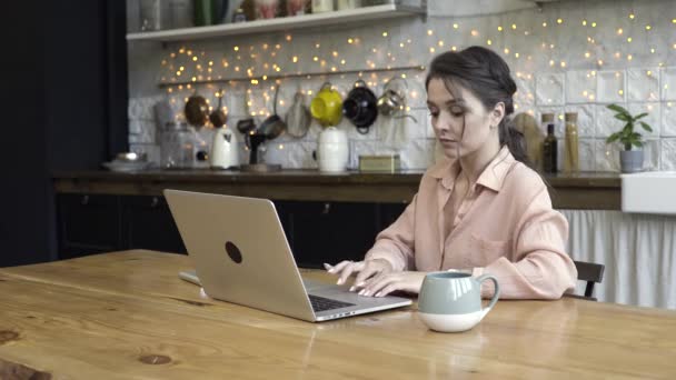 Mujer morena joven que trabaja en casa en el portátil en su cocina moderna. Imágenes de archivo. Hermosa mujer adulta buscando reflexivo y concentrado en su computadora portátil . — Vídeo de stock