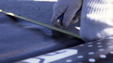 Siyah ruberoid tabakasını teknik kesici ile kesen beyaz koruyucu eldivenli işçi elleri için yakın çekim. Şarjör. İşçi, çatı malzemelerinden bir parça kesiyor..