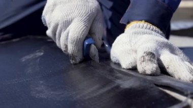 Siyah ruberoid tabakasını teknik kesici ile kesen beyaz koruyucu eldivenli işçi elleri için yakın çekim. Şarjör. İşçi, çatı malzemelerinden bir parça kesiyor..