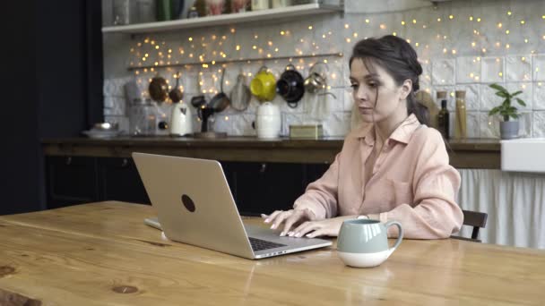 一个年轻的女人坐在厨房桌旁用笔记本电脑。 脸上挂着忧心忡忡的表情。 库存录像。 在现代设计的厨房工作的布鲁内特妇女. — 图库视频影像
